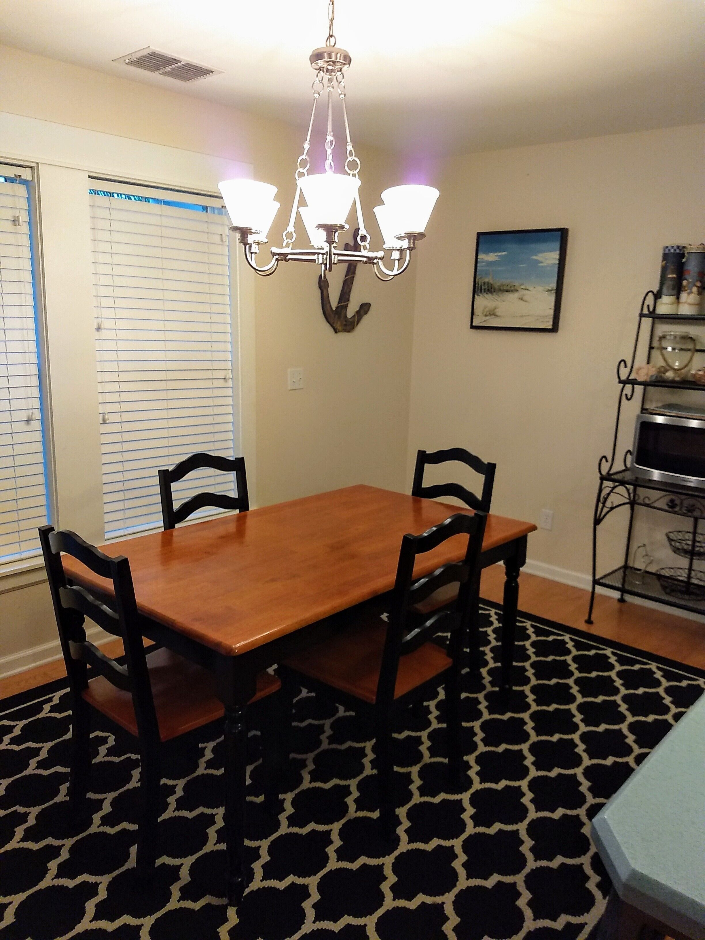 Un coin repas avec une table en bois et quatre chaises, un lustre, une décoration murale représentant une ancre, une fenêtre avec des stores et une étagère ornée d’objets décoratifs.