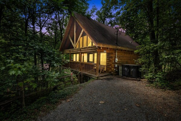 A log cabin with a wooden deck, a porch, and a gravel driveway surrounded by trees.