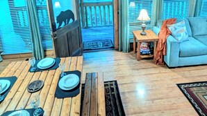 Een ruime kamer met een hoge plafond, houten balken, een eethoek met een tafel voor vier personen, een bank en uitzicht op bomen door grote ramen.