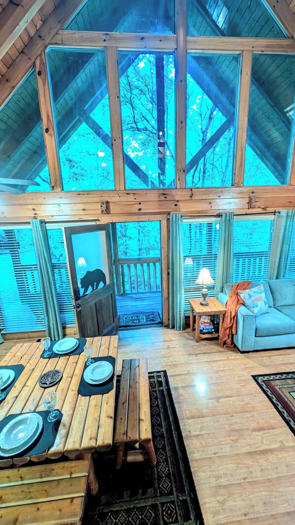 A spacious room with a high ceiling, wooden beams, a dining area with a table set for four, a sofa, and a view of trees through large windows. - Elegant Peaceful Mountain Cabin,  treehouse vibe, hot tub, fun games galore  (Pigeon Forge)