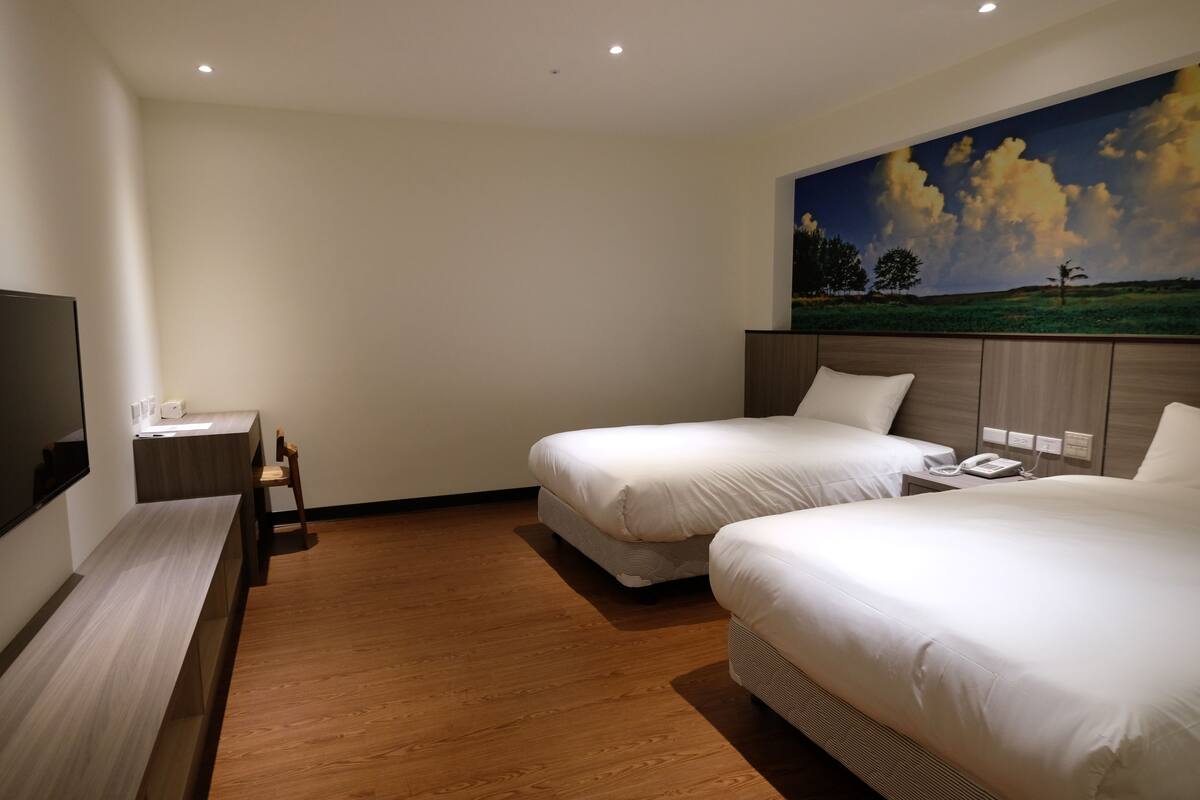 A hotel room with two beds, a desk, a chair, a TV, and a painting on the wall.