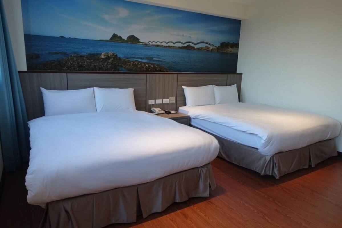 A hotel room with two beds, a wooden floor, and a large painting of a coastal scene on the wall.