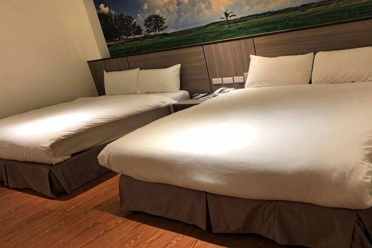 A hotel room with two beds, a wooden floor, a headboard with a landscape painting, and a nightstand with a lamp.