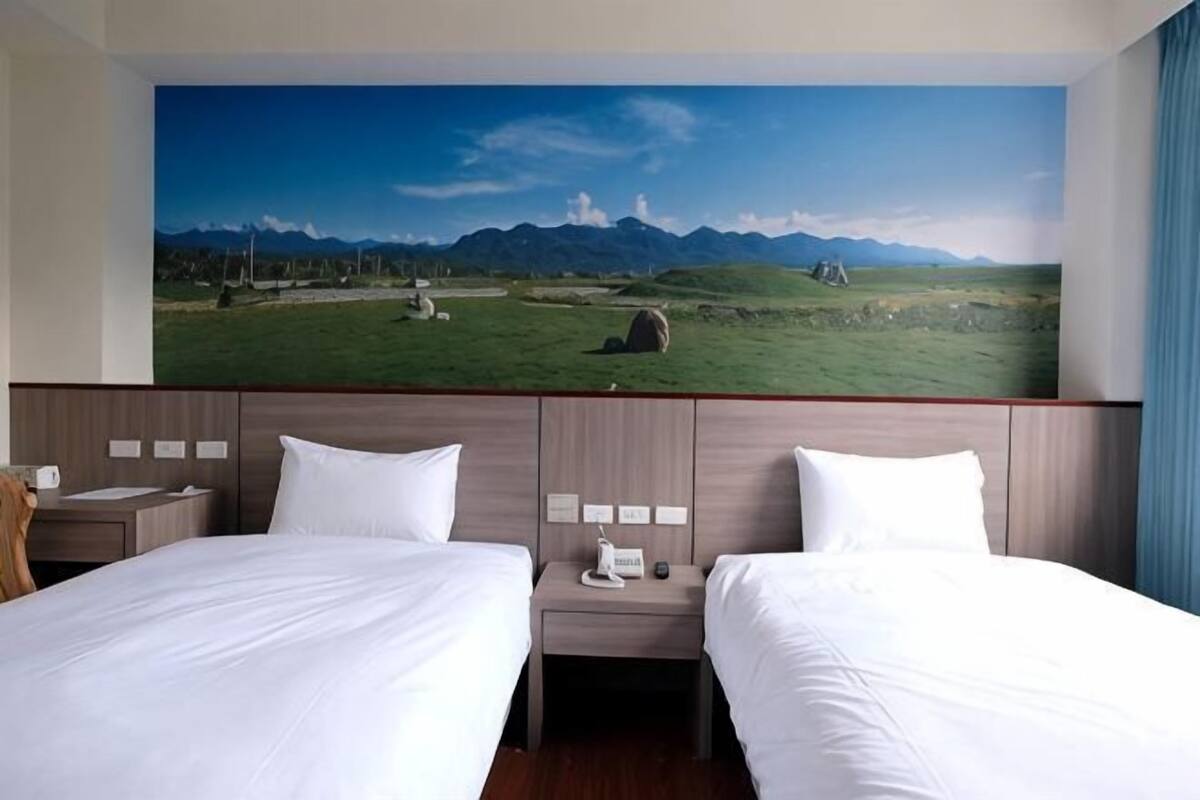 A hotel room with two beds, a large window, and a view of a landscape with mountains and grass.