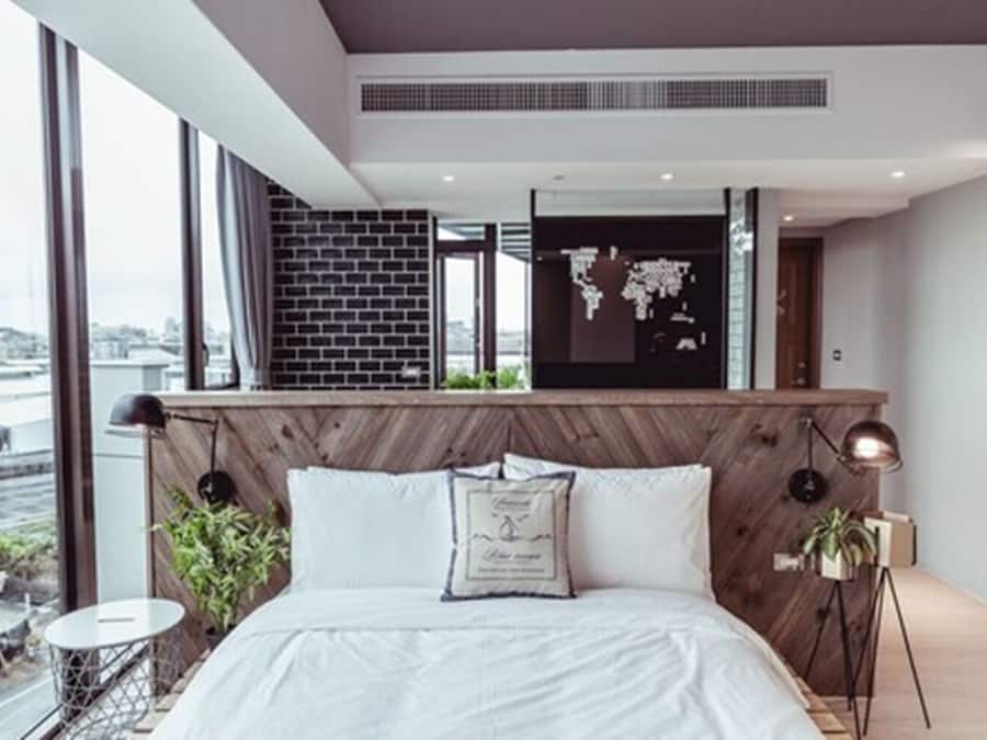 A modern bedroom with a large bed, a wooden headboard, a world map on the wall, and a view of the city through large windows.