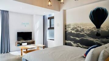 A modern hotel room with a large bed, a flat-screen TV, a desk, and a mural of clouds with a hot air balloon.