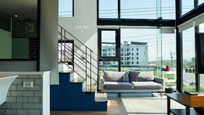 A modern interior with a staircase, a sofa, and large windows offering a view of buildings and a road.