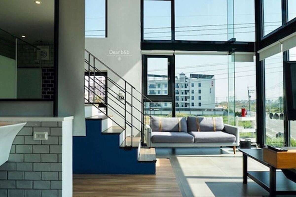 Een modern interieur met een trap, een bank en grote ramen met uitzicht op gebouwen en een weg.