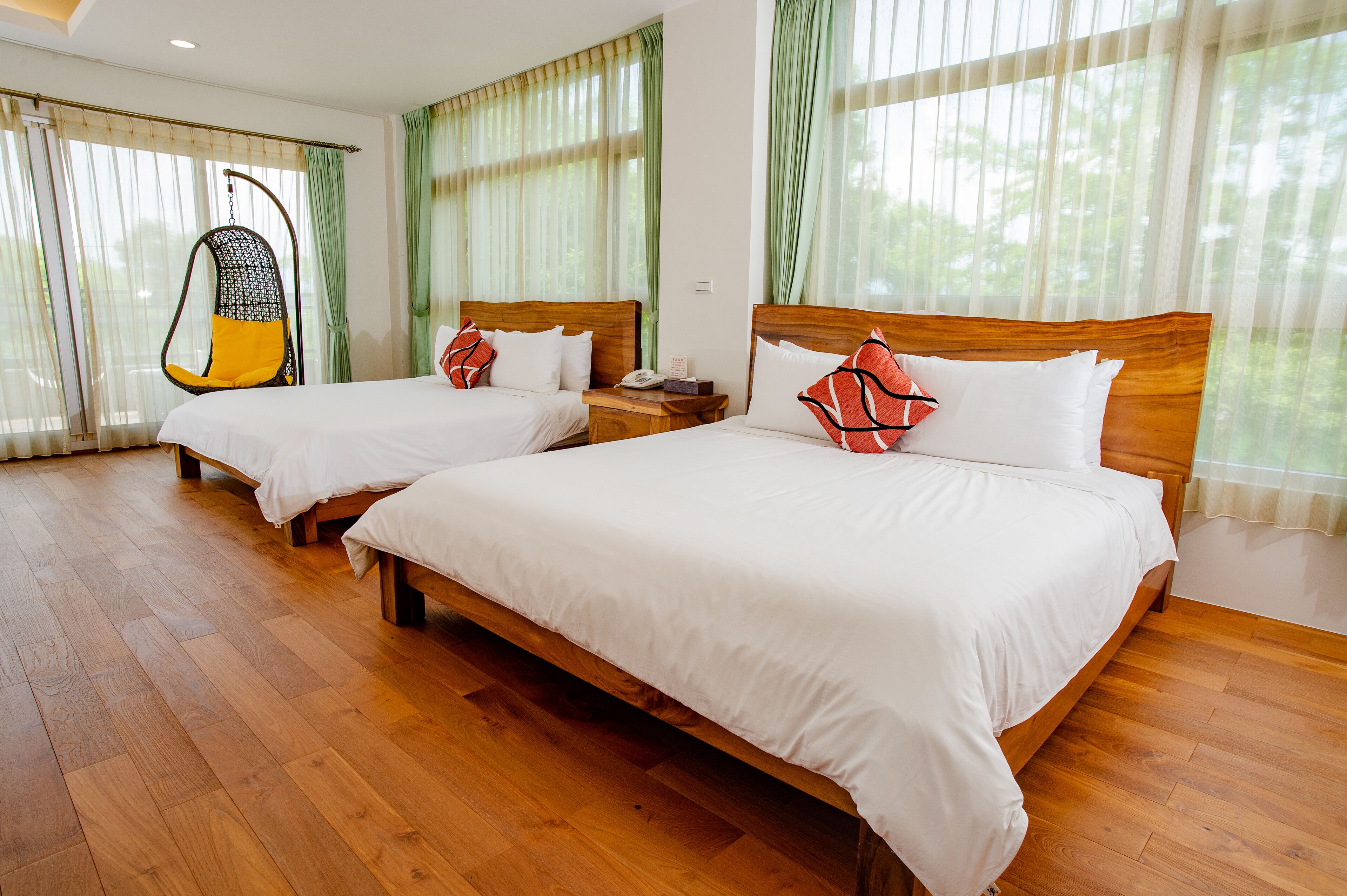 A hotel room with two wooden beds, a hanging chair, and large windows with sheer curtains.
