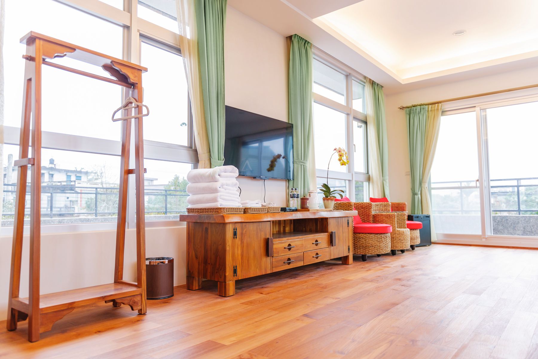 A spacious room with a wooden floor, a wooden cabinet with drawers, a television mounted on the wall, and a large window with green curtains.