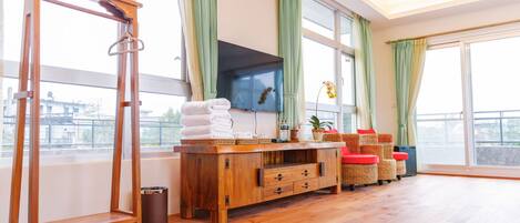 Una habitación amplia con suelo de madera, un mueble de madera con cajones, un televisor montado en la pared y un gran ventanal con cortinas verdes.