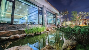 Un edificio moderno con grandes ventanales, fachada de piedra y un pequeño estanque con rocas y plantas.