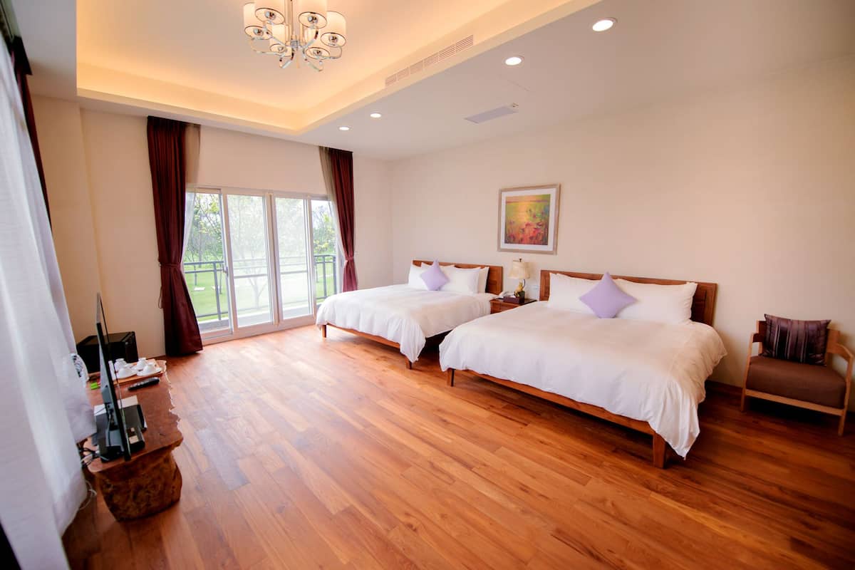 A hotel room with two beds, a wooden floor, a chandelier, a painting on the wall, and a balcony with curtains.