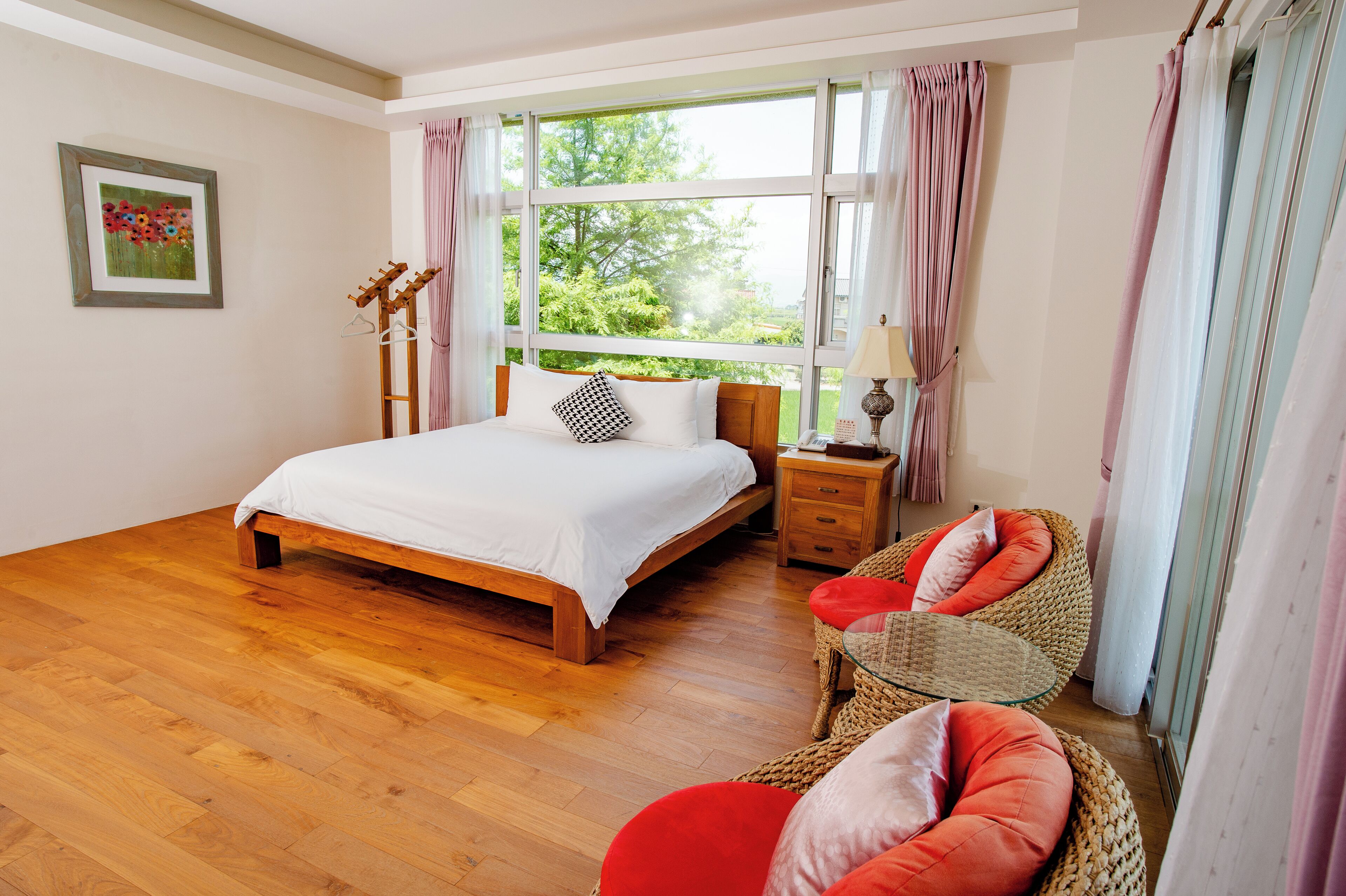 A bedroom with a wooden bed, a wicker chair with red cushions, a bedside table with a lamp, and a window with curtains.