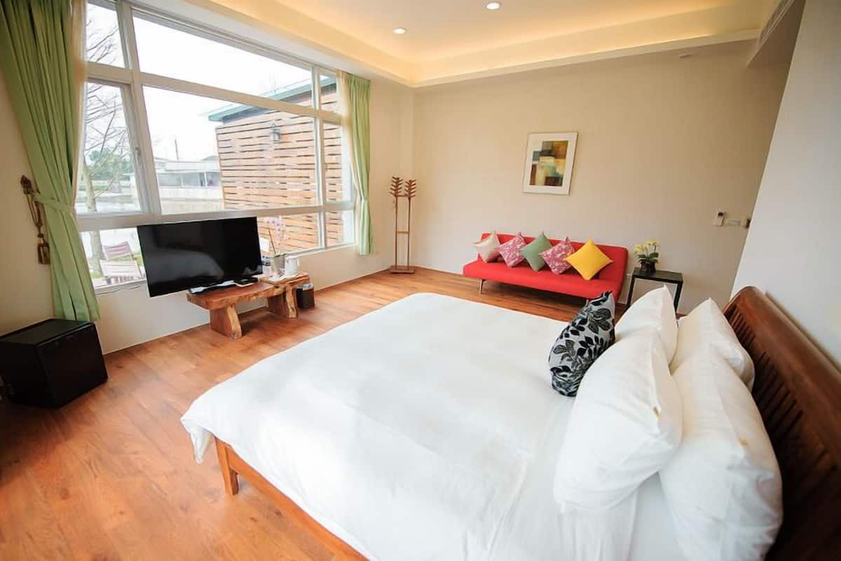 A bedroom with a large bed, a TV on a wooden stand, a red sofa, and a window with curtains.