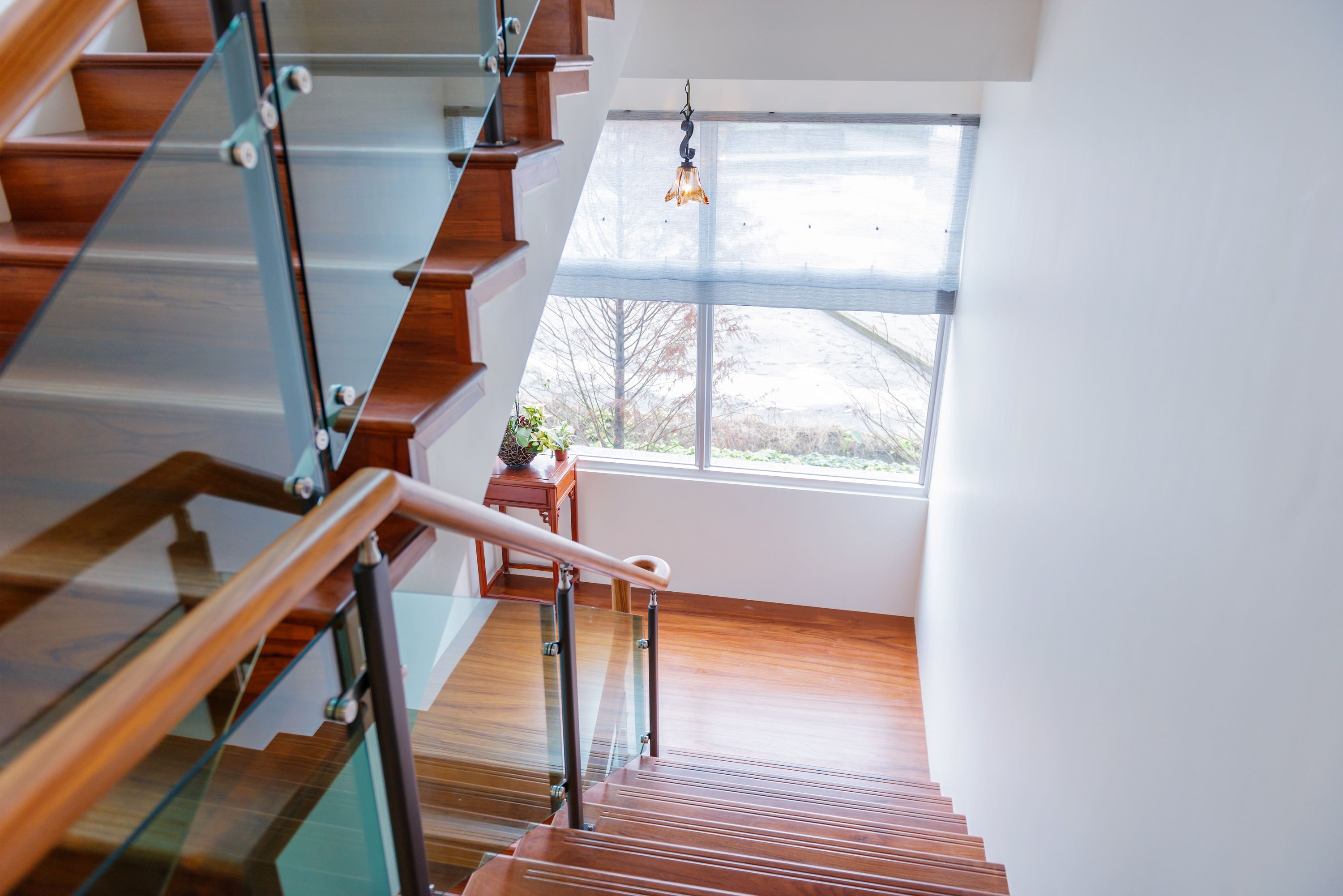 A modern staircase with glass railings and wooden steps, leading to a room with a large window and a hanging light fixture.