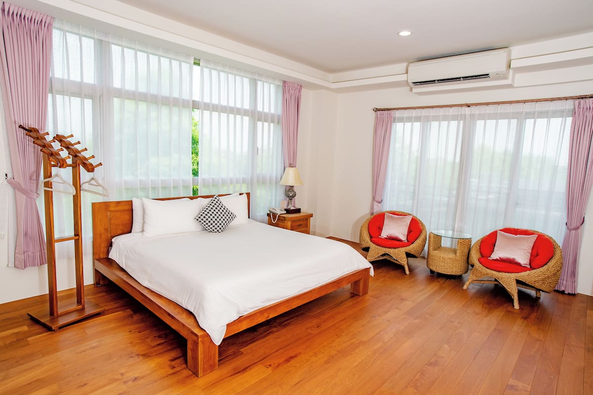 A bedroom with a wooden bed, a bedside table, a lamp, two wicker chairs, and large windows with sheer curtains.