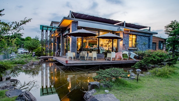 Una casa moderna con una terraza que da a un estanque, con mobiliario exterior y un jardín bien cuidado.
