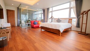 Una habitación de hotel moderna con suelo de madera, una cama con ropa blanca, un sofá rojo con cojines de colores, un separador de vidrio y un ventanal con cortinas.