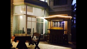 An outdoor patio area with a wooden table and chairs, a bar counter, and a large window with curtains. - Bann Dao (Lam Luk Ka)