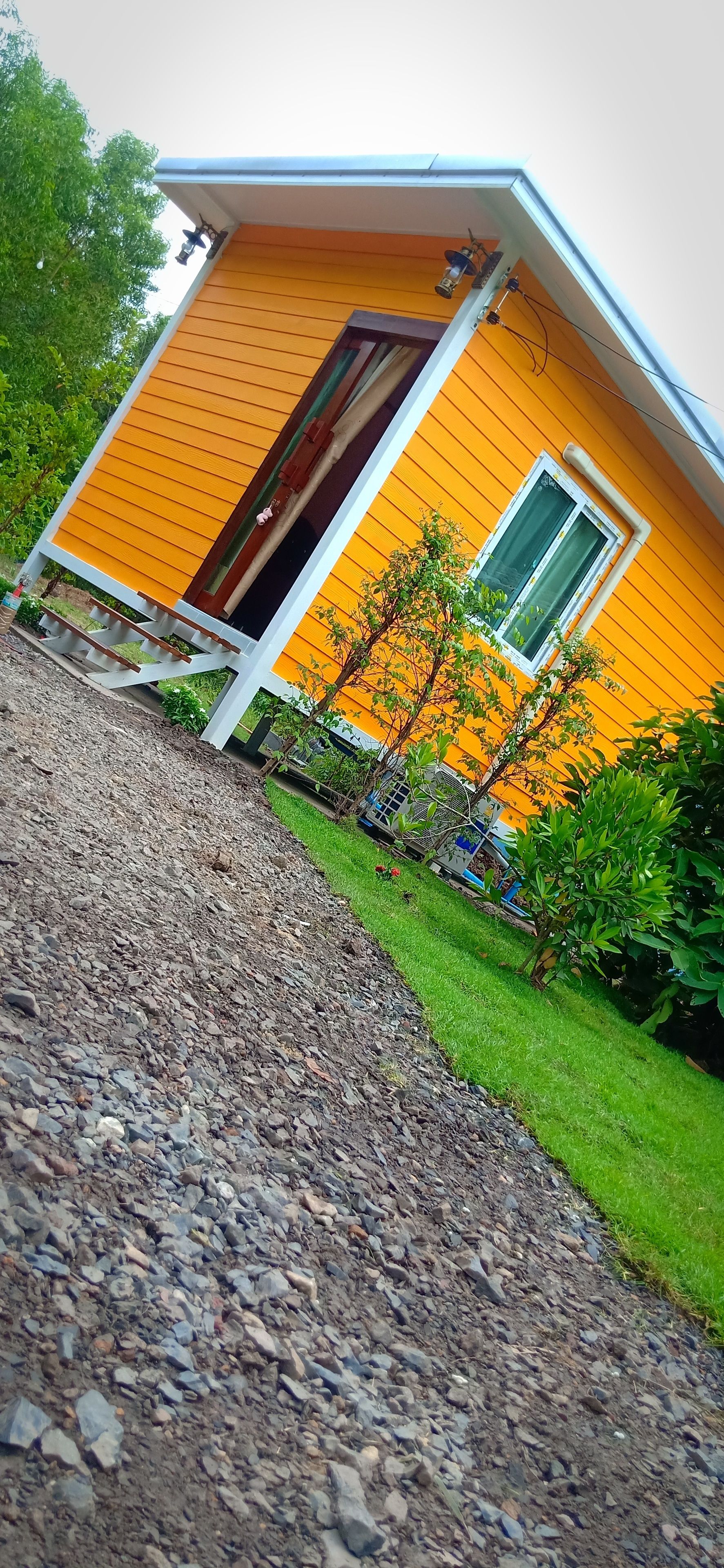A yellow building with a wooden staircase leading to a door, surrounded by greenery and a gravel path.