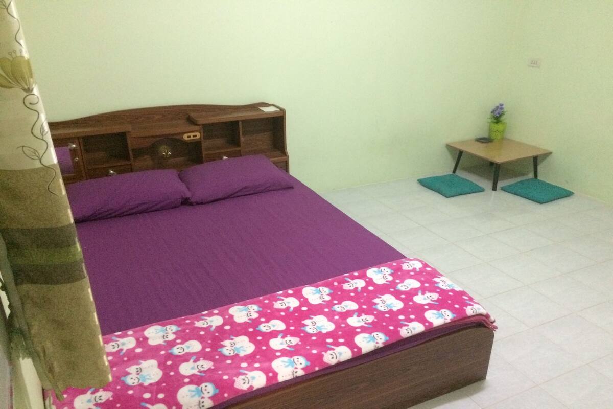 A bedroom with a bed, a nightstand, a small table with a vase, and a curtain.