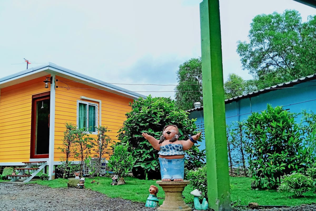A small yellow house with a red door, surrounded by greenery and a blue building in the background.
