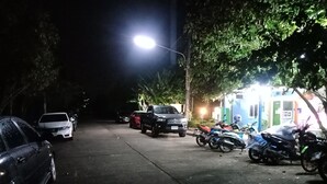 A nighttime parking area with cars and motorcycles, a brightly lit street lamp, and a building with a blue and green facade. - Suansuppachok Inn (Ongkharak)
