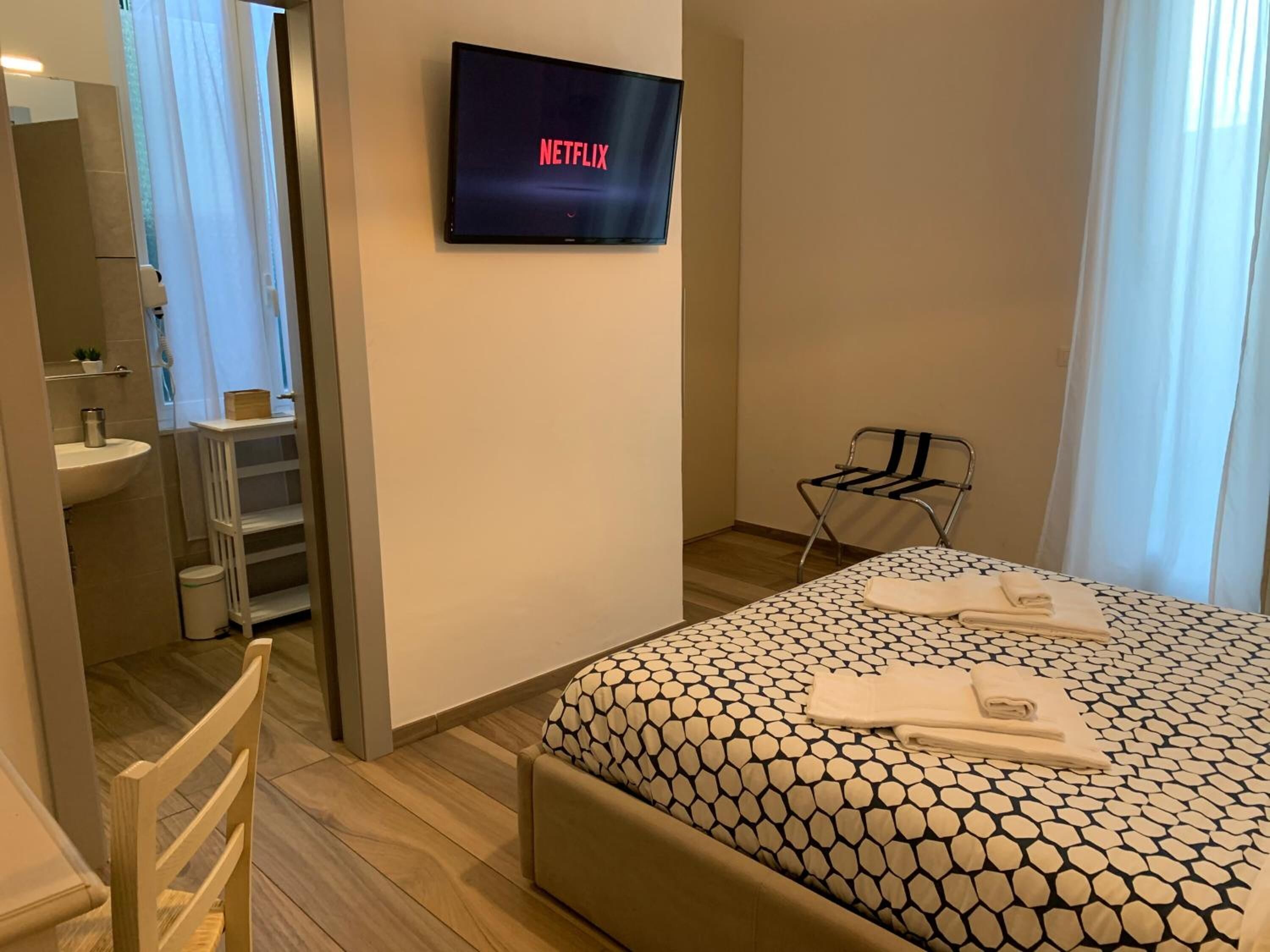Habitación de hotel con cama, un televisor mostrando 'NETFLIX', baño con lavamanos y espejo, y una silla.