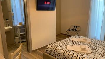 Habitación de hotel con cama, un televisor mostrando 'NETFLIX', baño con lavamanos y espejo, y una silla.