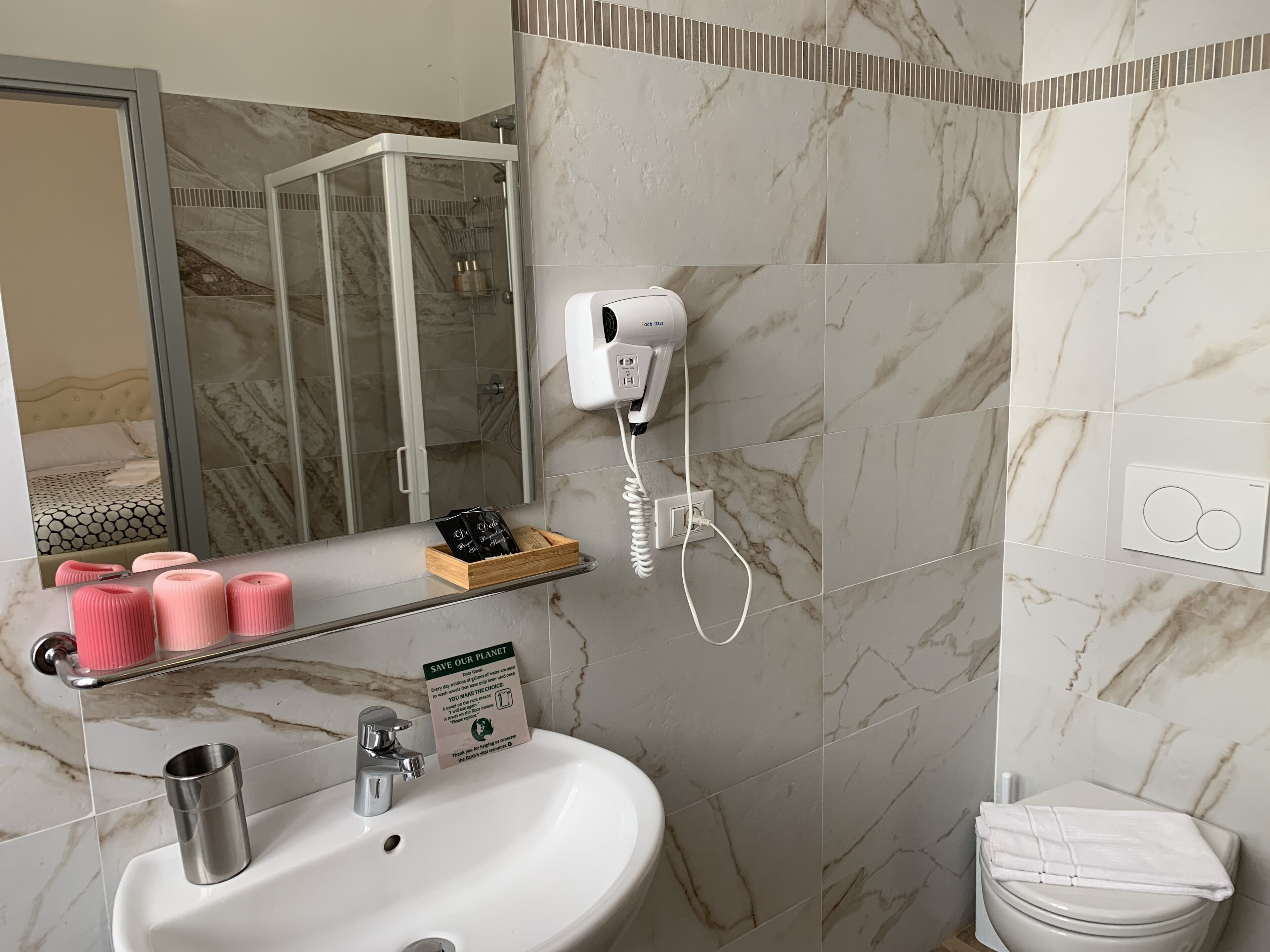 Ein Badezimmer mit einer marmorverkleideten Wand, einer Duschkabine aus Glas, einem weißen Waschbecken mit Chromarmatur, einem Föhn an einer Wandsteckdose und einem Handtuch auf der Toilette.