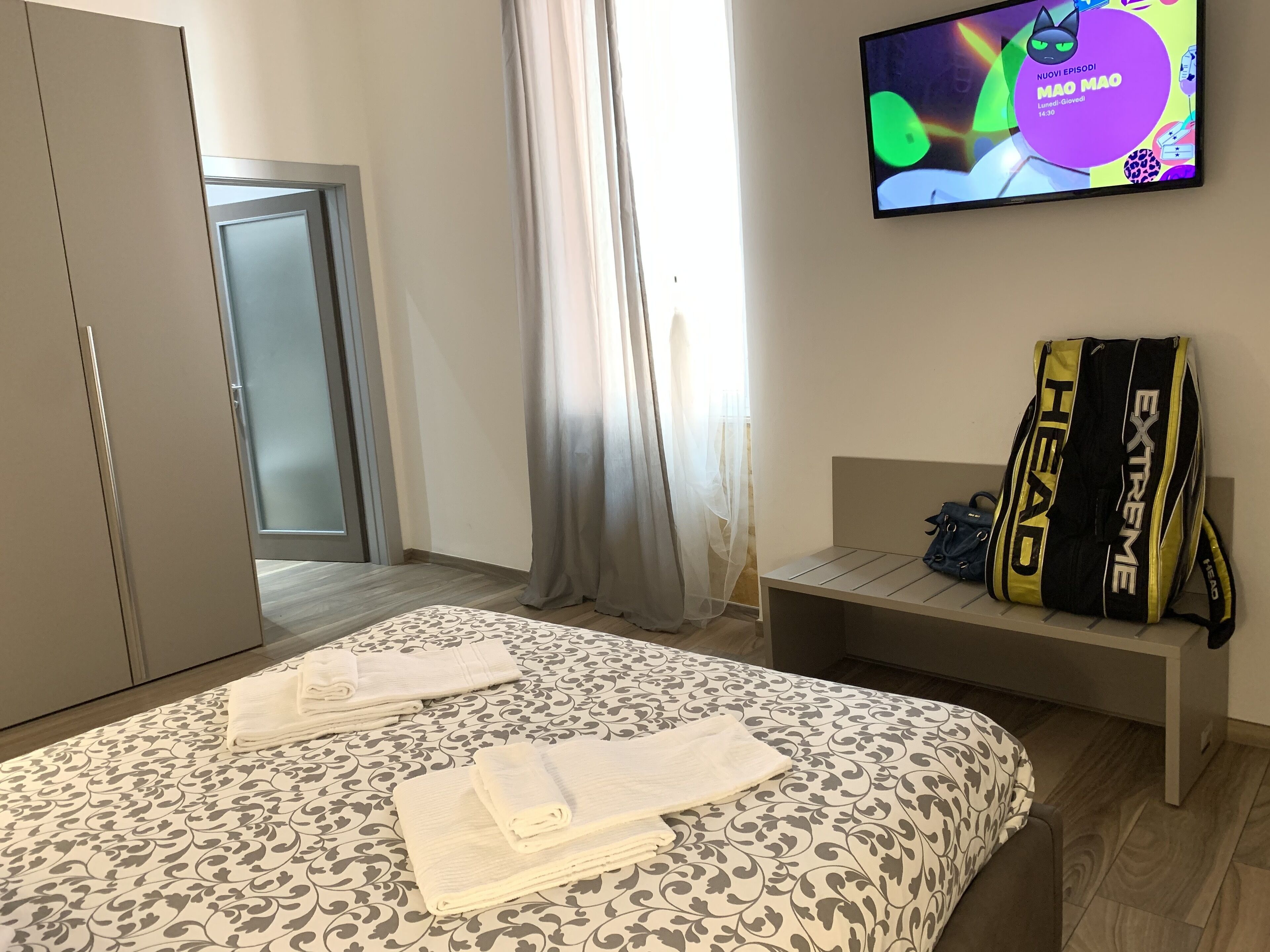 Quarto de hotel com cama, televisão exibindo um desenho animado, guarda-roupa e um banco com uma mochila.