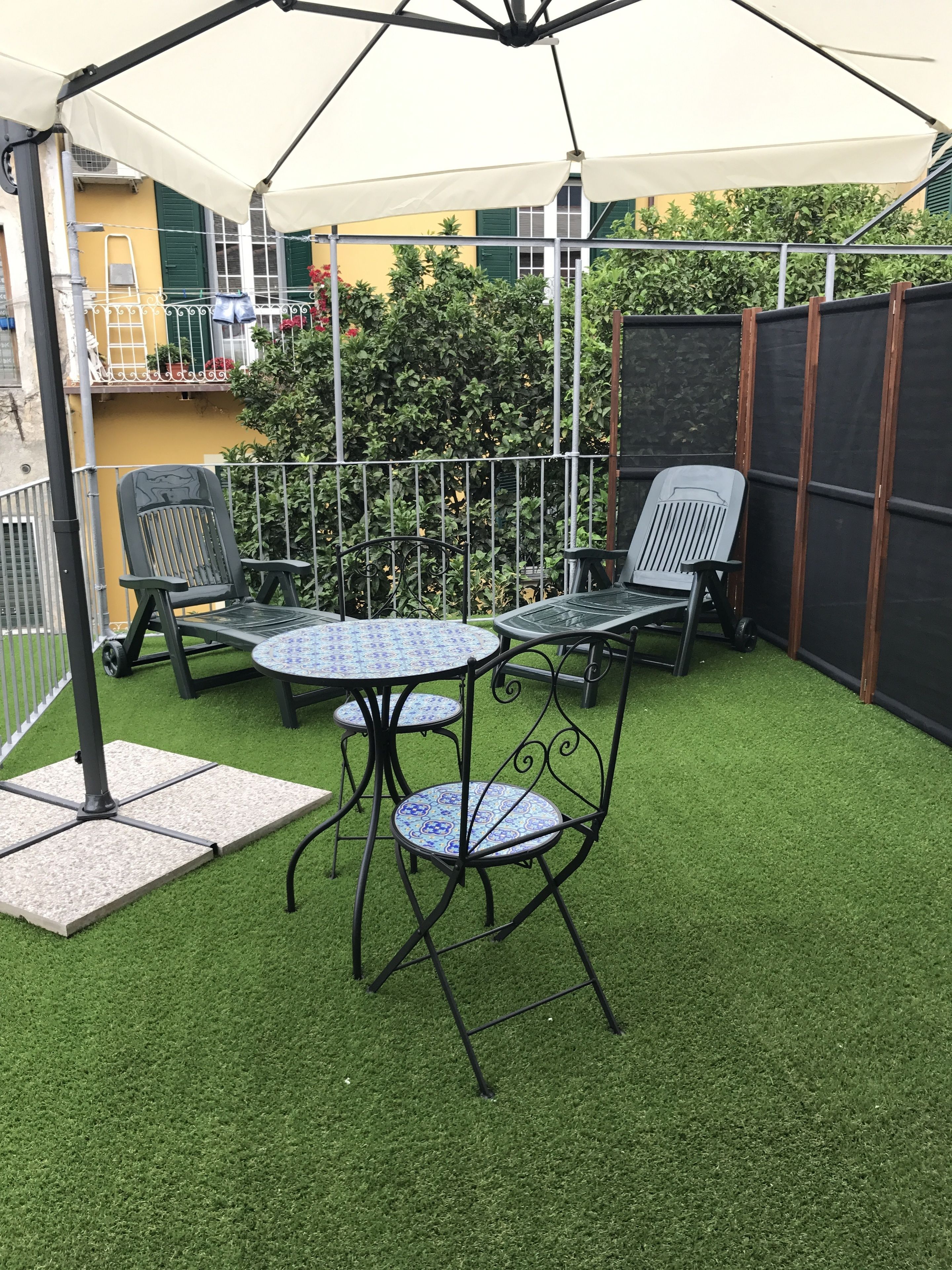 Uma área de pátio com uma mesa redonda e duas cadeiras, cercada por uma grade de metal e um arbusto verde.