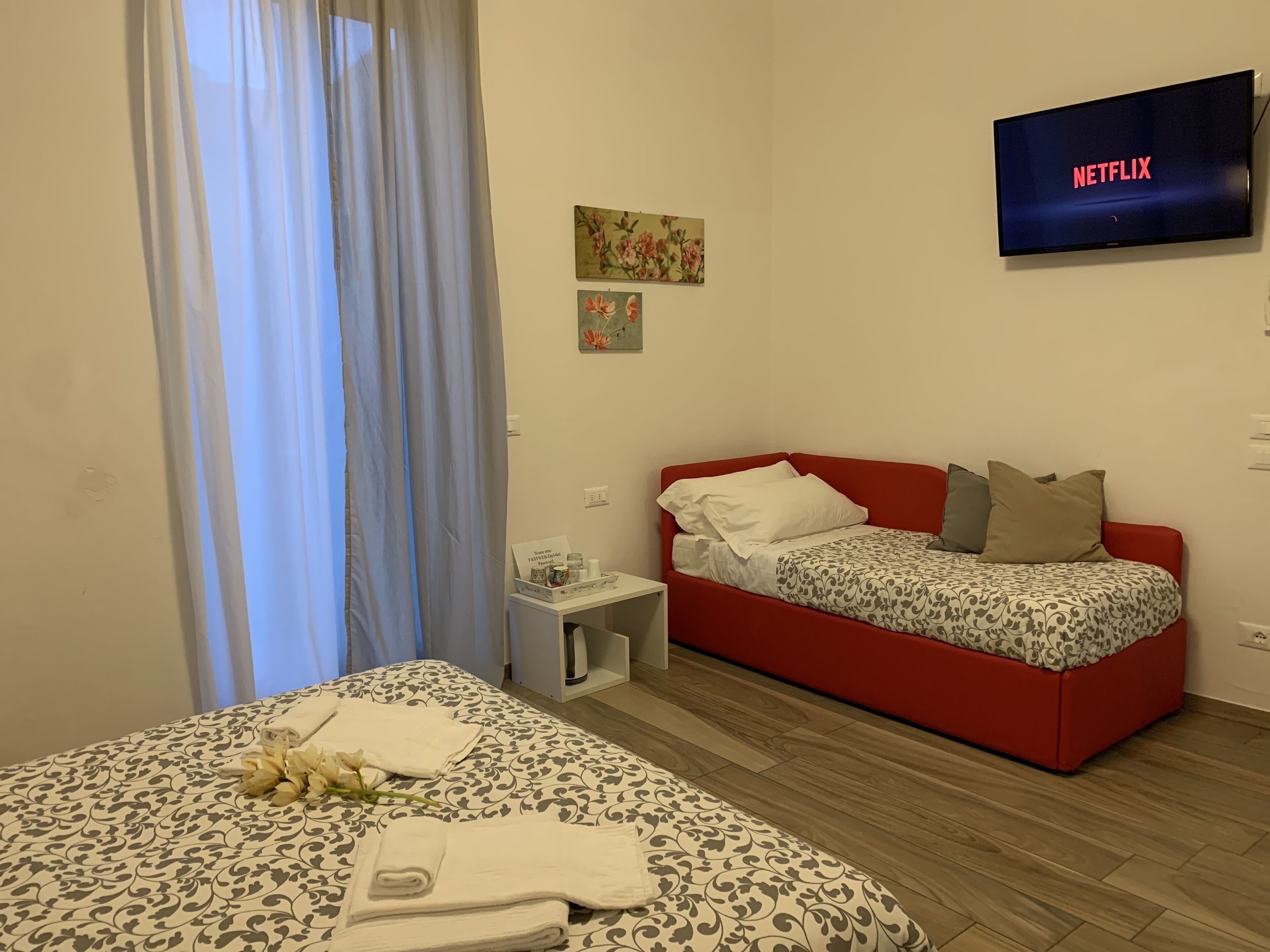 Een hotelkamer met een bed, een rode slaapbank, een flatscreen-tv waarop 'NETFLIX' staat, een klein tafeltje met een dienblad en een raam met gordijnen.