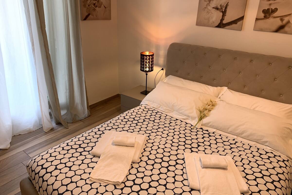 A neatly made bed with a geometric patterned bedspread, a bedside lamp, and a vase with flowers.
