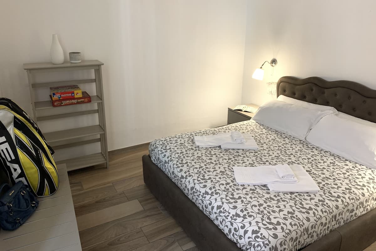 A neatly made bed with a patterned bedspread and pillows, a bedside lamp, a nightstand with a book, a shelf with boxes, and a bag on the floor.