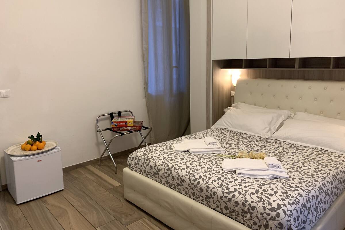 A hotel room with a bed, a small white refrigerator, a chair, and a window with curtains.