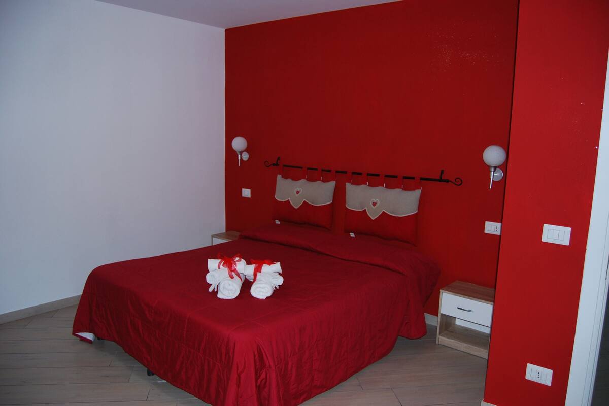 Une chambre avec une couvre-lit rouge, une petite table de chevet et un luminaire fixé au mur.
