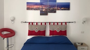 Een slaapkamer met uitzicht op de stad, een bed met blauw beddengoed, een rode stoel en een nachtkastje met een lamp.