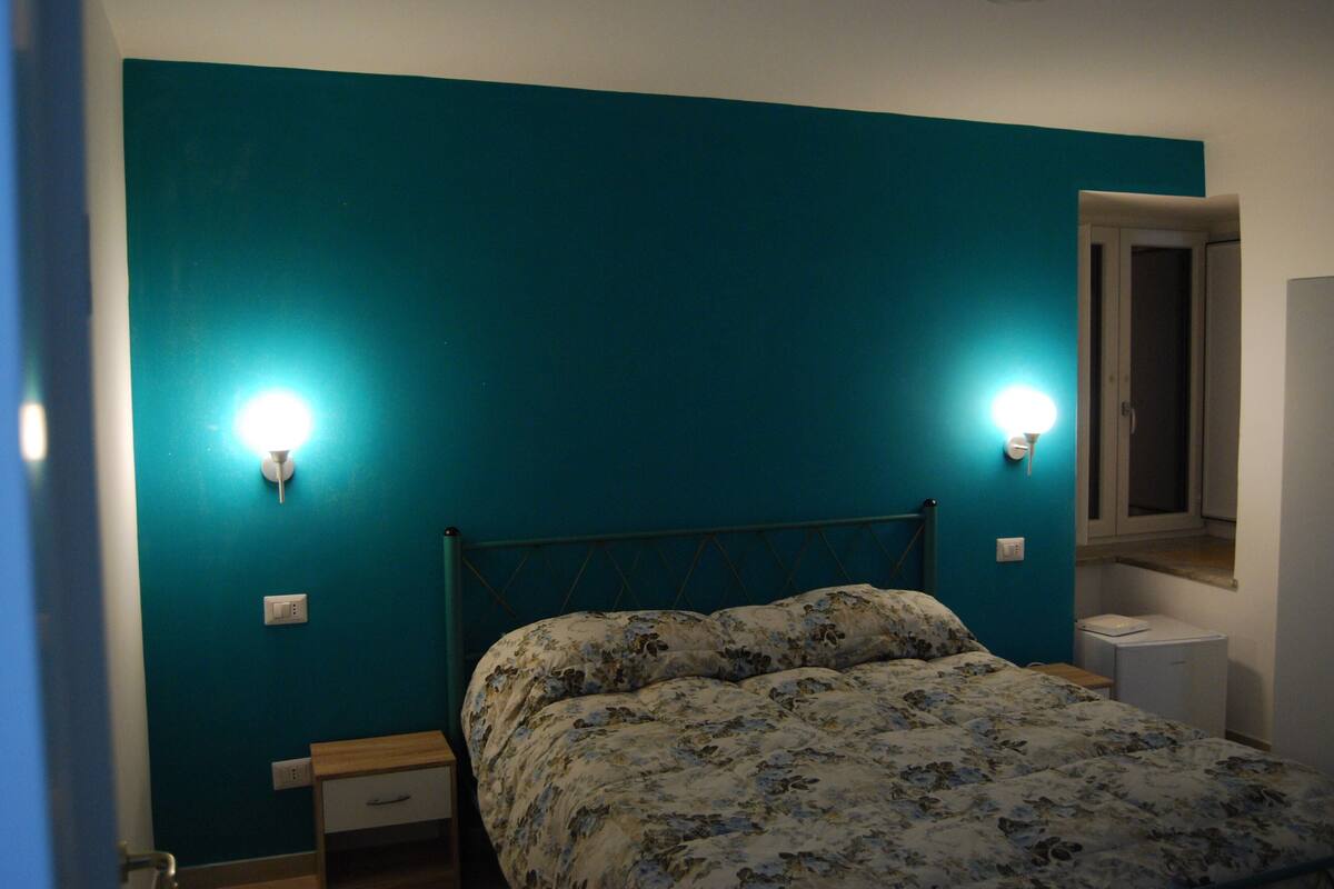 Une chambre avec des murs de couleur turquoise, un lit recouvert d’une couette à motifs floraux, deux lampes de chevet, une table de chevet et une petite table d’appoint.