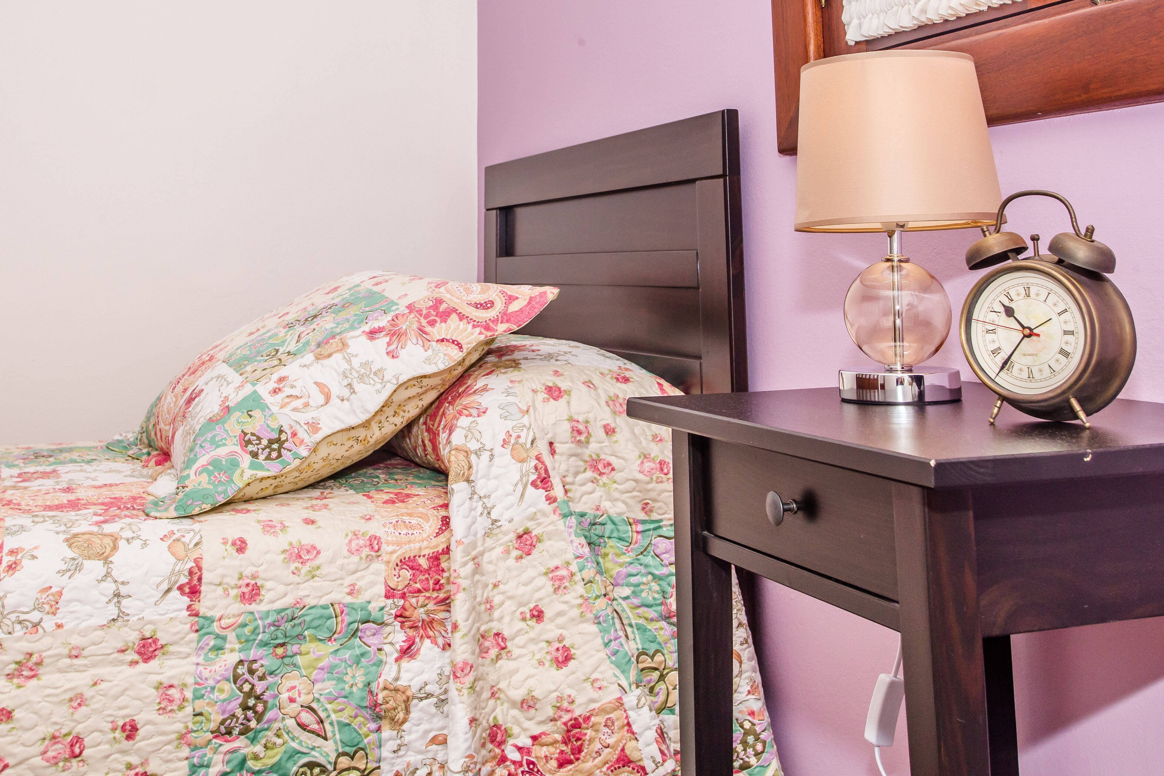 A neatly made bed with floral bedding, a bedside lamp, and an alarm clock on a wooden nightstand.