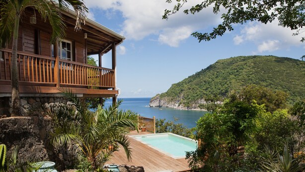 A wooden house with a balcony overlooking a clear blue sea and a lush green hill.