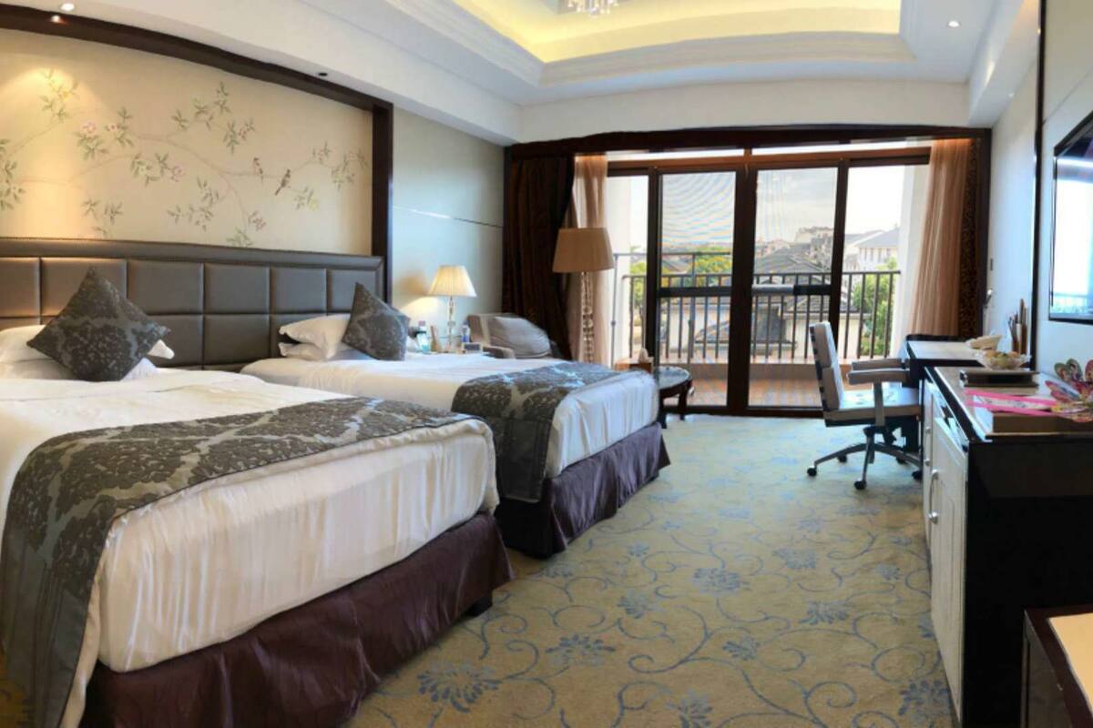 A hotel room with two beds, a desk, a chair, and a balcony with a view.