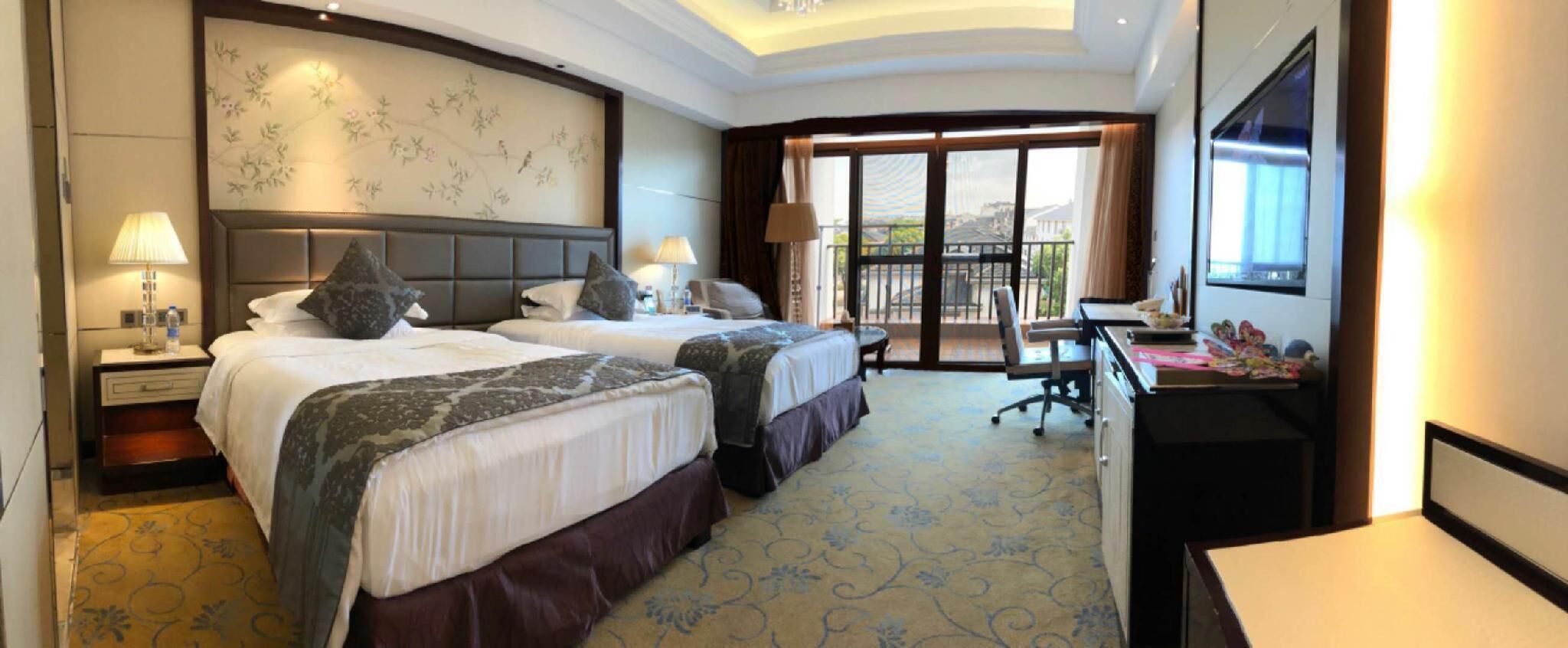 A hotel room with two beds, a desk, a chair, a balcony with a view, and a large window.
