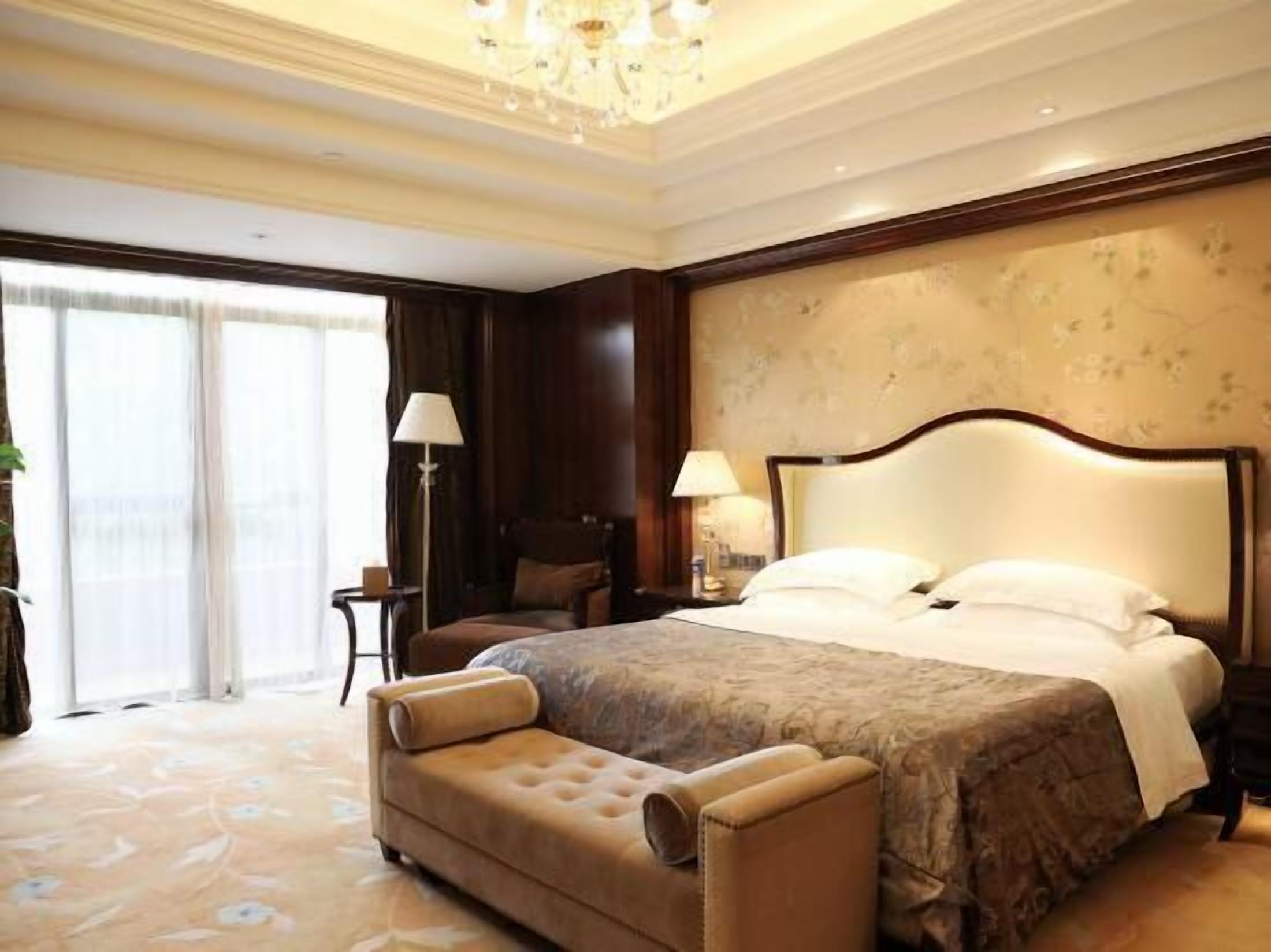A hotel room with a large bed, a chaise lounge, a chandelier, and a view of the outdoors.