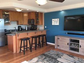 A modern kitchen with wooden cabinets, a granite countertop, a built-in microwave, and a large flat-screen TV. - Ocean And Mountain Views (Kihei)