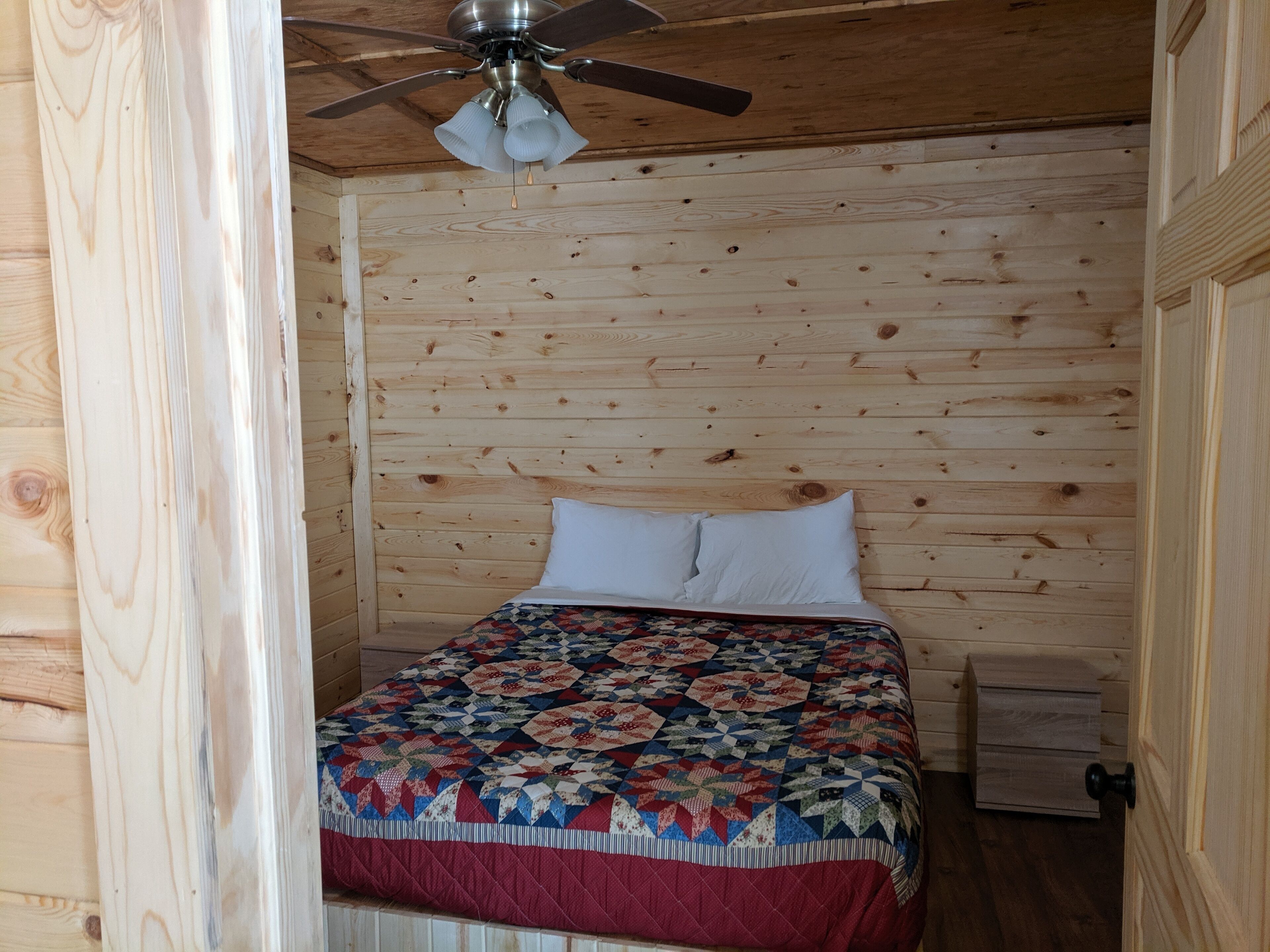 Una habitación con paneles de madera, una cama, un ventilador de techo y una mesita de noche.