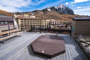A deck with picnic tables and a hot tub, overlooking a mountainous landscape with buildings in the background. - Snowcrest Condo, short walk from lifts (Crested Butte)
