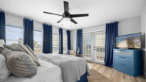 A bedroom with a bed, a ceiling fan, a TV, and a view of the beach through the window.