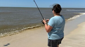 Eine Person beim Angeln an einem sandigen Strand, im Hintergrund das Meer.
